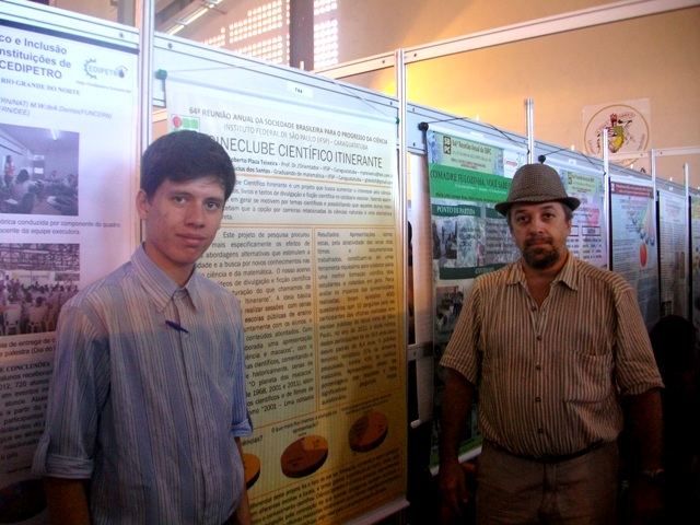 O aluno Gabriel Lúcius dos Santos e o professor Ricardo Plaza posam em frente ao pôster explicativo do Cineclube Científico Itinerante